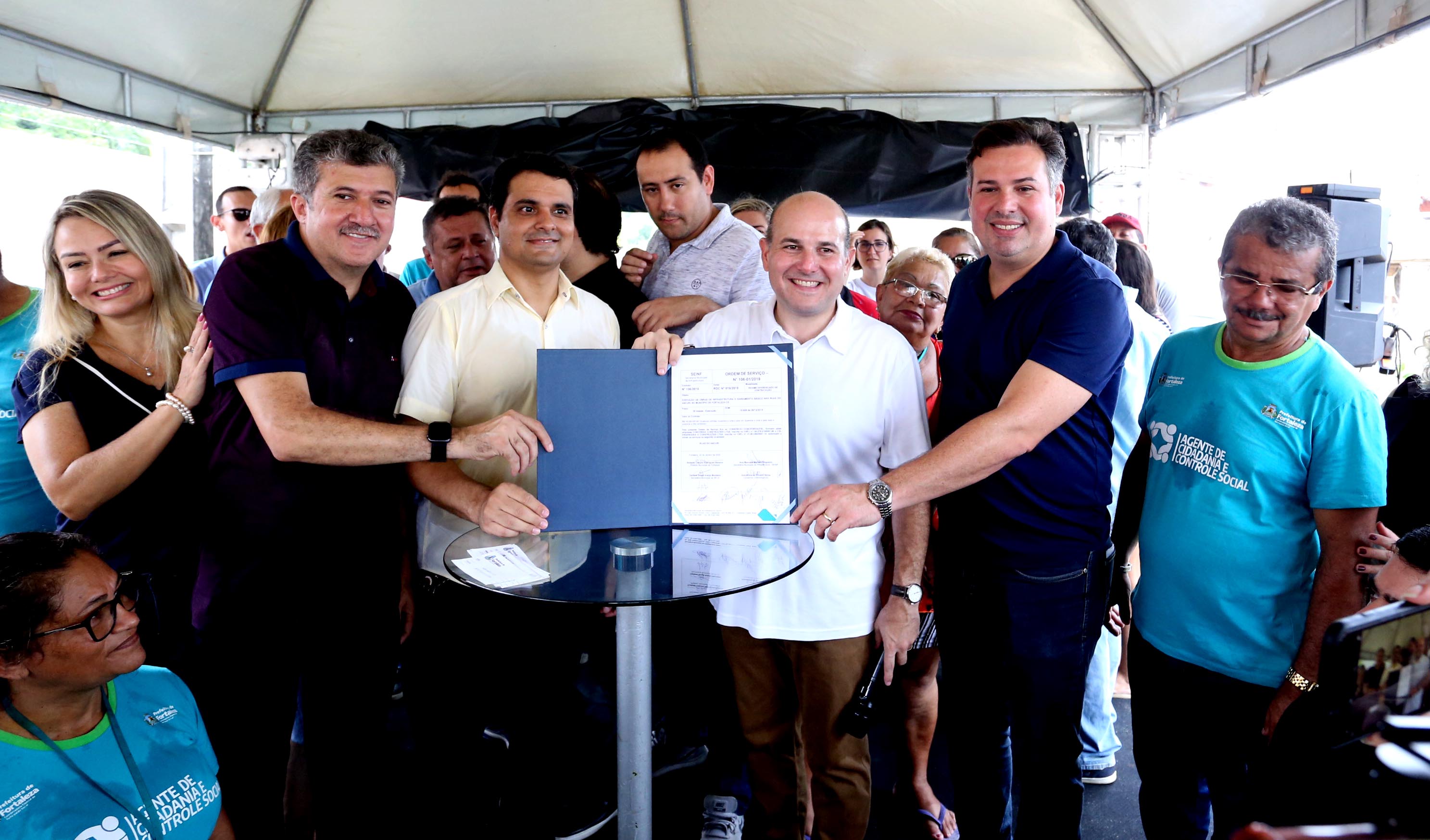 prefeito em cima de palco ao lado de pessoas segurando documento e sorrindo para foto